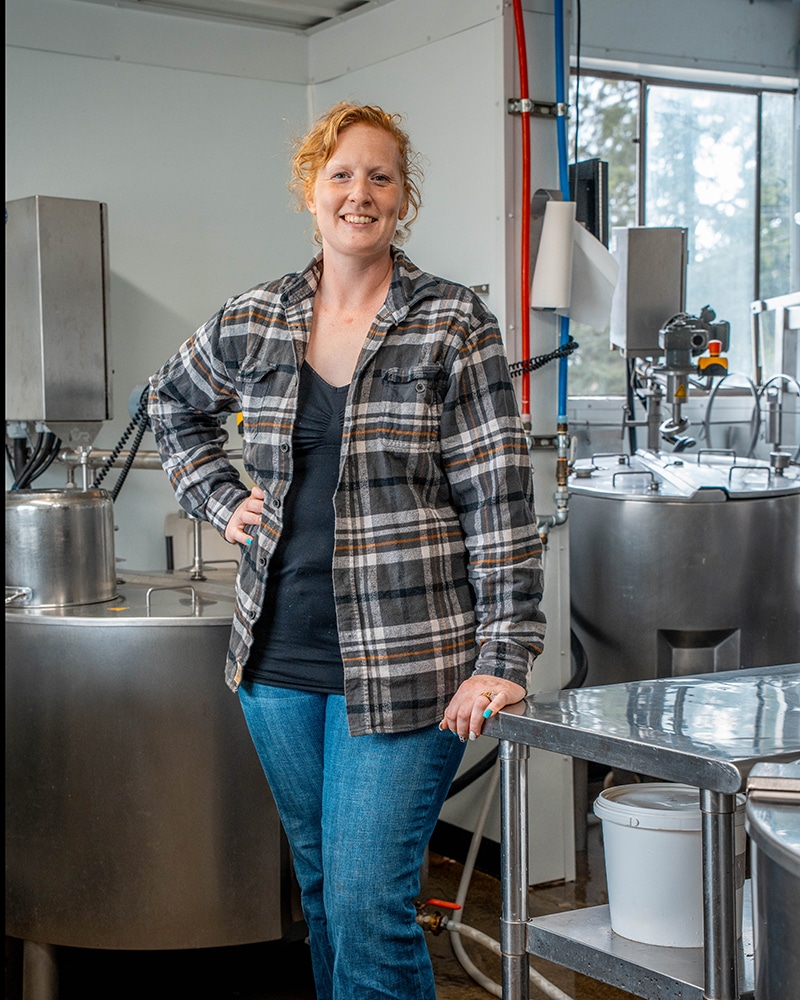 A woman in a flannel shirt and bluejeans stands in the middle of a creamery room with metal cheese vats and other equipment.